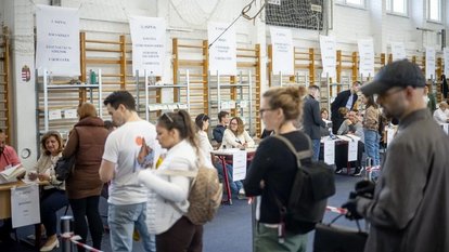 People queue indoors as election staff process ballots at multiple tables.