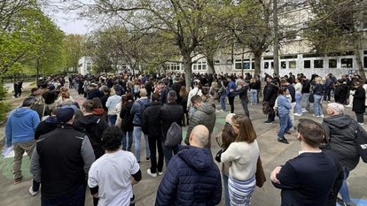 A large crowd waits along a tree-lined walkway near the entrance to a polling station.