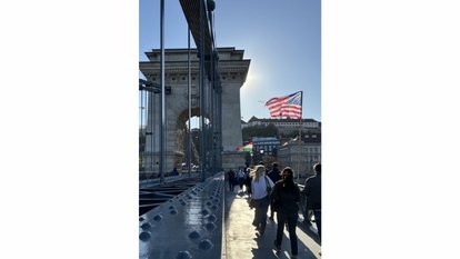 People walk along Budapest’s Chain Bridge toward a stone tower, with the bridge’s steel structure in the foreground and a large U.S. flag waving in the sunlight above the far end.