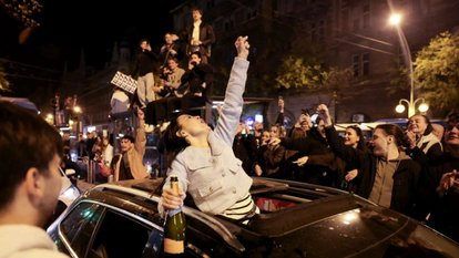 A cheering crowd gathers under streetlights; many film on their phones.