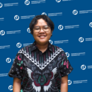 A photo of Dhea Ramadhani--sporting short curly hair, glasses, and black batik shirt--with FNF blue banner as the background.