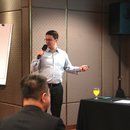 Dr. Stefan Diederich, holding a mic, in front of the room. On his left is a blank flipboard. On his right is a black table with glasses on top.